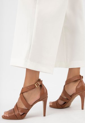 Pieds portant des sandales à talons hauts marron avec des brides et boucles autour de la cheville, associées à un pantalon blanc large et court.