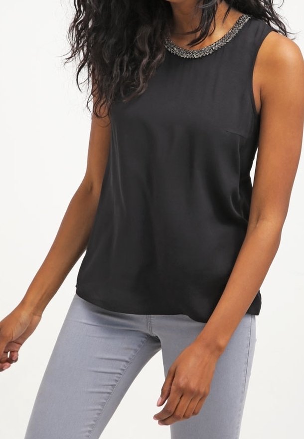 Femme portant un top noir sans manches avec un col orné de perles et un jean gris clair ajusté sur fond blanc