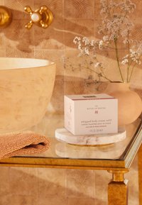 Vit fyrkantig låda märkt "whipped body cream refill" på en marmorbrygga, med en texturerad beige handduk och en gyllene handfat i ett badrum i neutrala toner.