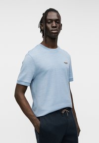 Ανοιχτό μπλε κοντομάνικο πουκάμισο από υφή ύφασμα με στρογγυλή λαιμόκοψη και μικρό λογότυπο στο στήθος.