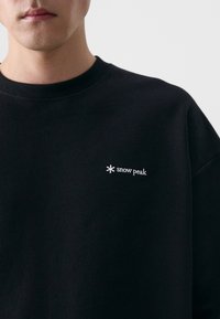 Sweatshirt noir avec un col rond, des poignets côtelés et un logo « snow peak » brodé en blanc sur la poitrine gauche. Texture lisse.