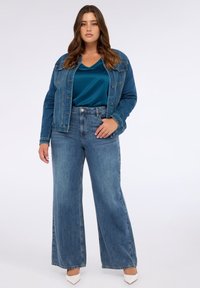 Džínová bunda a kalhoty s širokými nohavicemi v světle modré, doplněné saténovým topem v tyrkysové. Hranaté bílé podpatky dotvářejí outfit. Jsou viditelné hladké textury.