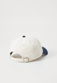 White corduroy baseball cap with a navy bill, adjustable strap at the back, and a matching navy top button. Textured fabric and structured shape.