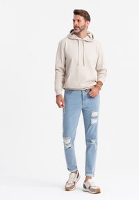 Czapka w kolorze jasnego beżu z kapturem, z przednią kieszenią, w połączeniu z jasnoniebieskimi dżinsami z przetarciami i licznymi dziurami, oraz sportowymi butami w neutralnych odcieniach.