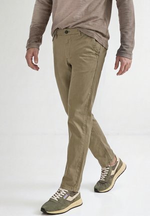 Homme portant un pantalon beige, des baskets vertes et beiges, et une chemise marron à manches longues marchant sur un fond blanc.