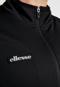 Schwarze Zip-Jacke aus strukturiertem Material mit Stehkragen. Hervorstechendes weißes ellesse-Logo auf einem schwarzen Patch. Sichtbares Reißverschlussdetail.