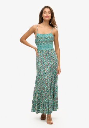 Vestido maxi floral con un cuerpo fruncido, tirantes finos, patrones vibrantes de flores azules y rojas sobre un fondo verde, complementado por un dobladillo con volantes.