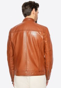 Giubbotto di pelle marrone con texture liscia, design strutturato e polsini con zip. Cuciture dettagliate lungo la schiena e le spalle.