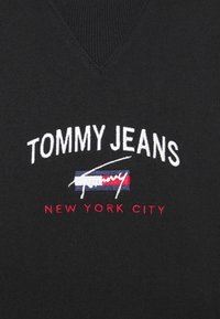 Tissu noir avec le texte blanc "TOMMY JEANS" au-dessus d'un petit drapeau rouge, blanc et bleu, et le texte rouge "NEW YORK CITY" brodé en dessous.