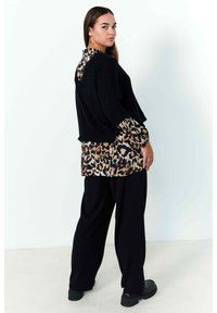 Pull en maille noir avec une chemise à imprimé léopard en dessous, pantalons large noirs et chaussures noires imposantes. Le mannequin pose de profil contre un fond clair.