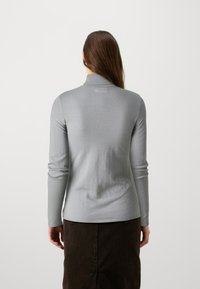 Haut gris à manches longues avec un col montant, texture côtelée et coupe ajustée. Porté avec une jupe brun foncé, montrant une silhouette simple et épurée.