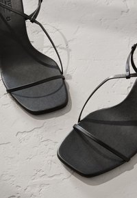 Sandales à talons noires avec fines brides croisées. Design à bout carré et texture lisse et mate sur la semelle intérieure. Esthétique minimaliste.