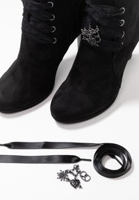 Bottines en suède noir avec des lacets ruban ornés de breloques métalliques ; lacets ruban noirs supplémentaires et breloques posés à côté des bottines sur une surface blanche.