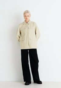 Beige langermet jakke med to brystlommer, knappelukking foran og klassisk krage, paret med svarte vide bukser og støvler.