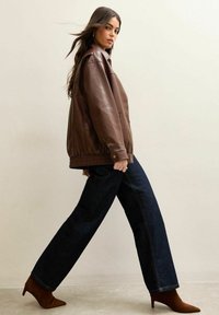 Giacca in pelle marrone con orlo a coste, abbinata a jeans a gamba larga blu scuro e stivaletti marroni con punta appuntita; texture liscia e design strutturato.