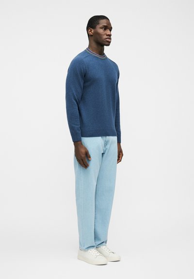 Homme portant un pull bleu avec un col multicolore, un jean bleu clair et des baskets blanches, debout devant un fond clair uni.