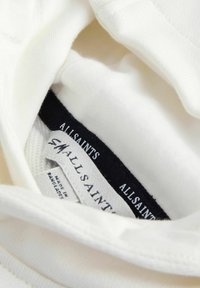 Vêtements blancs avec des étiquettes de marque visibles indiquant « ALLSAINTS » et « SMALL », incluant une étiquette « Fabriqué au Bangladesh » à l'intérieur du vêtement.