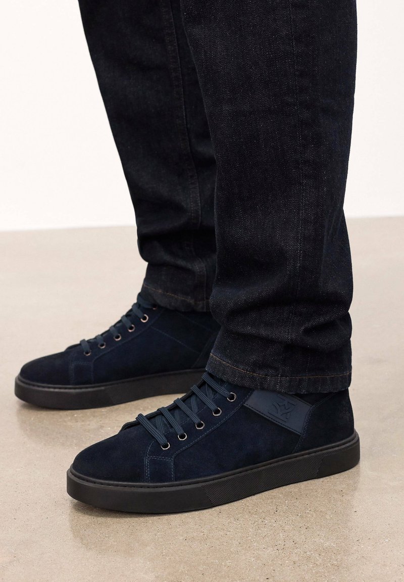 Navy zila zamšas augstie sporta apavi ar tonāliem šņorēm, gumijas zolēm un sānu akcentu paneli. Valkāti ar tumšas džinsa biksēm uz gludas virsmas.