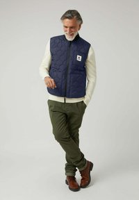 Chaleco acolchado azul marino con escote redondo, cremallera frontal y dos bolsillos; combinado con un suéter crema y pantalones verde oliva; botas marrones con cordones.