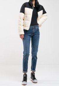 Valkoinen ja musta puhvitakki, jossa on korkea kaulus ja tikkikuvio, yhdistettynä sinisiin farkkusiin ja mustiin korkeakorkoisiin tennareihin.