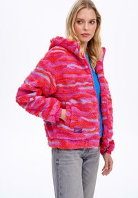 Kolorowa polarowa kurtka z kapturem w odcieniach różu i fioletu, ozdobiona falistymi wzorami, posiada pełny zamek błyskawiczny, kaptur oraz elastyczne mankiety. Teksturowany materiał.
