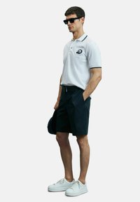 Witte poloshirt met marineblauwe accenten, marineblauwe shorts en witte sneakers. Het shirt heeft een logo op de borst; de shorts hebben zijzakken.