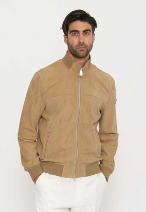 Uomo con capelli scuri e barba che indossa una giacca color cammello in pelle scamosciata con zip e pantaloni bianchi, in piedi contro uno sfondo chiaro e semplice, con le mani in tasca.