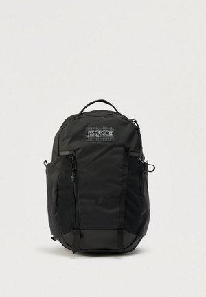 Sac à dos Jansport noir avec poignée supérieure, poche zippée avant, poches latérales et logo de la marque visible sur un fond uni.