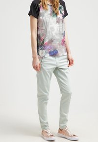 Tričko s krátkým rukávem s barevným tropickým potiskem a černými rukávy, spárované s mentolově zelenými slim-fit kalhotami a světle růžovými teniskami.