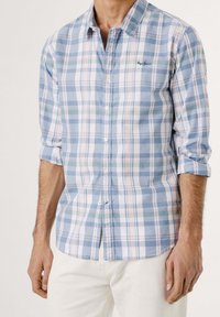 Muž v kostkované košili s modro-bílo-růžovým vzorem, s vyhrnutými rukávy, v kombinaci s bílými kalhotami, stojící na jednobarevném pozadí.