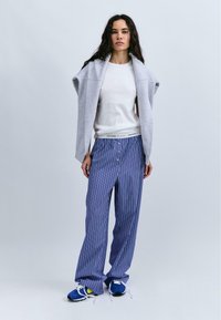 Sweatshirt gris surdimensionné porté par-dessus un t-shirt blanc, associé à un pantalon large rayé bleu et des chaussures de sport bleues avec des lacets blancs.