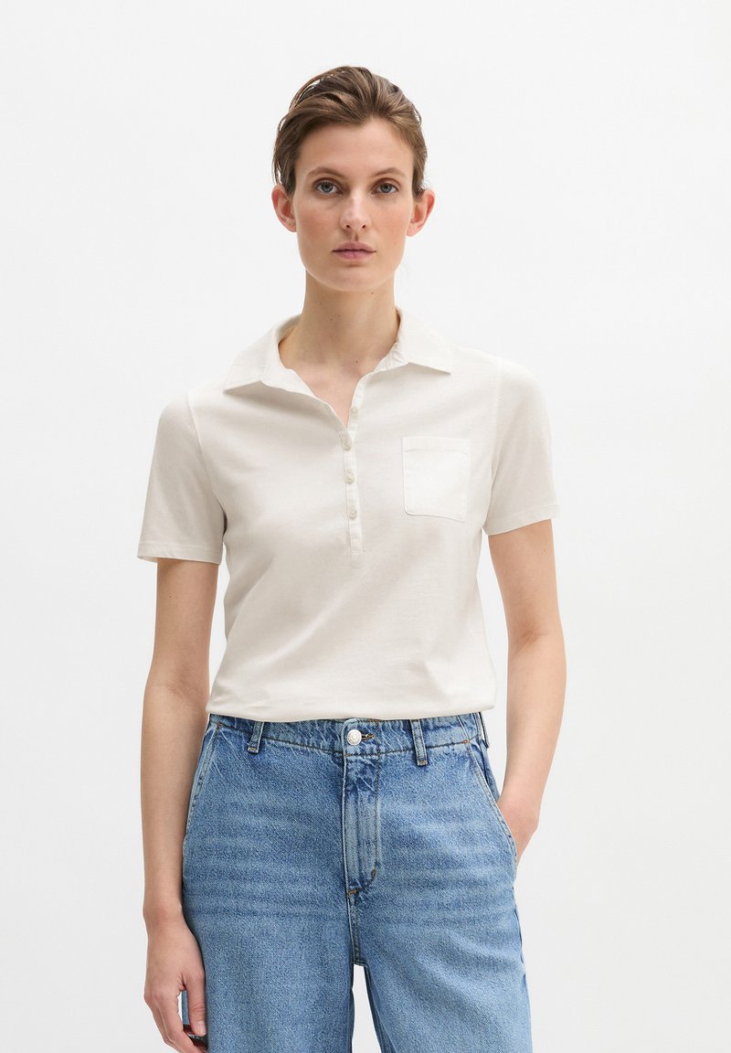 Frau mit kurzen braunen Haaren, die ein weißes, kurzärmeliges Hemd mit Kragen und Knöpfen sowie eine blaue, hoch taillierte Jeans trägt, steht mit einer Hand in der Tasche.