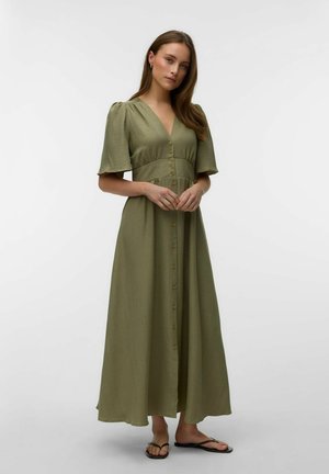Kvinde iført olivengrøn maxikjole med knapper foran, korte pufærmer og sorte flade sandaler, stående mod en ensfarvet lys baggrund.