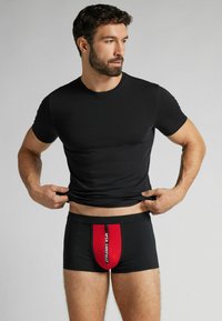 Czarny sportowy t-shirt z krótkim rękawem, zestawiony z czarnymi bokserkami z czerwonym panelem na przodzie z zamkiem, opatrzonym napisem "OTWIERAJ OSTROŻNIE". Gładka tekstura materiału.