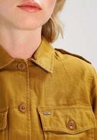 Senapsgul skjorta med knappar, två framfickor, bruna knappar och en liten "OBEY"-etikett på den högra fickan, tillverkad av strukturerad tyg.