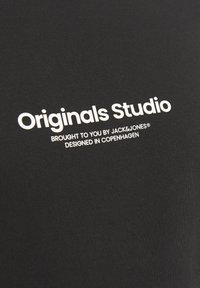 Černá tkanina s hladkou texturou, na které je bílý potisk s textem: "Originals Studio," "přináší vám JACK & JONES," a "navrženo v Kodani."