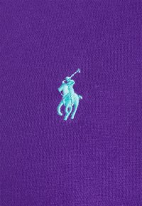 Blå broderad polospelare med klubba på lila tygbakgrund.