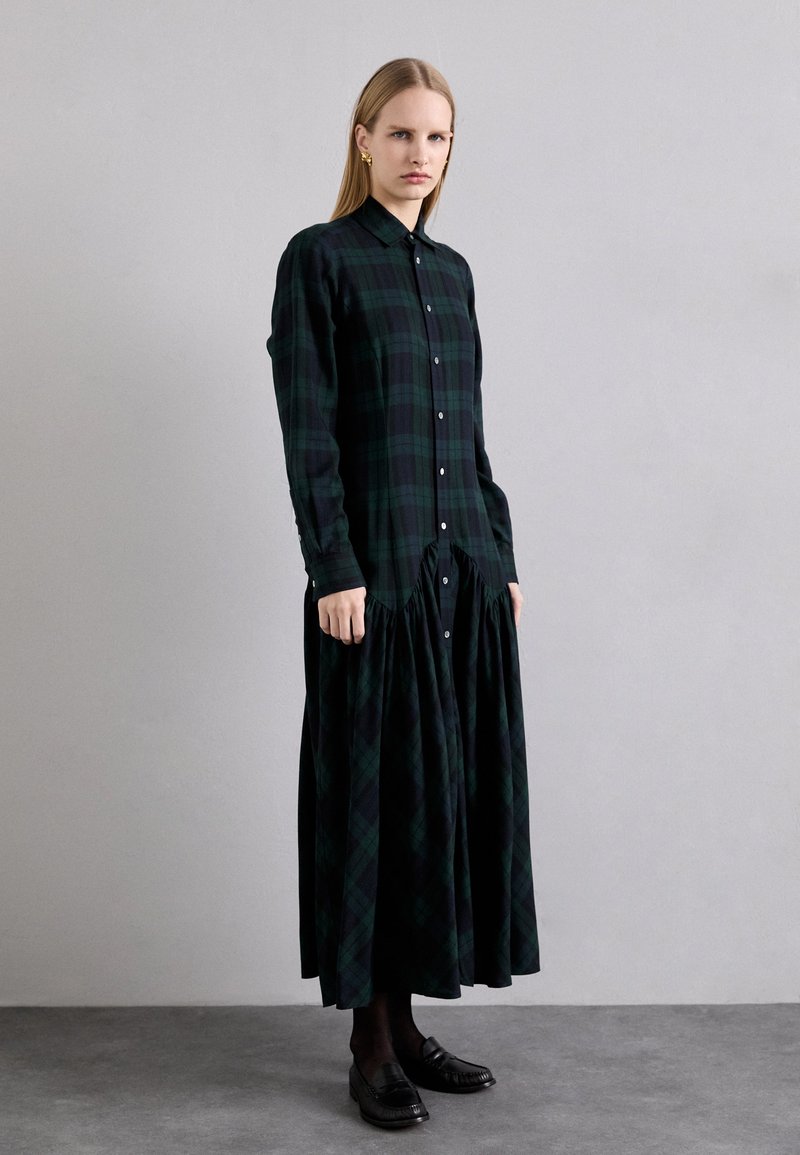 Long, button-up dress made of dark green and black plaid fabric. Features a gathered waist and long sleeves. Paired with black loafers.