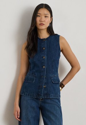 Hosszú sötét hajú nő, aki ujjatlan, gombos sötét farmermellényt és hozzáillő farmert visel, világos fal előtt áll, kezét a csípőjén tartva.