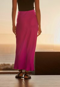 Lunga gonna rosa realizzata in tessuto leggero, aderente in vita con una silhouette fluida. Indossata con sandali neri con cinturini.