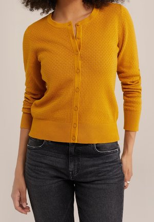 Modèle portant un cardigan texturé jaune moutarde, boutonné sur le devant, et un jean foncé, sur un fond beige uni.