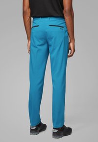 Pánské modré kalhoty z hladké látky, s kapsami na zadní straně zdobenými černými detaily a střih na míru. Nošeny s černými botami.