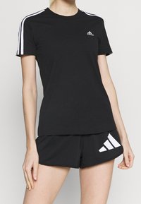 Svart bomullst-shirt med korta ärmar, vita axelstrimmor och en liten vit Adidas-logga på bröstet, kombinerad med svarta shorts.