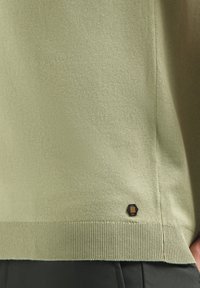 Lichtgroene trui van zachte stof, met een geribde onderrand en een hexagonaal label met strepen nabij de linksonder.