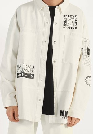 Chemise blanche à boutons avec texte graphique noir et motifs, portée sur une chemise noire, avec des mots comme "READY," "OUTPUT," et "RAN."