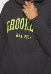 Sudadera negra con capucha y bolsillo tipo canguro en el frente, con un texto en amarillo verdoso llamativo que dice "BROOKLYN USA 2007". Tela suave y capucha con cordón ajustable.