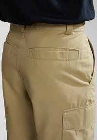 Pantalones cargo color caqui con un bolsillo plano en la pierna derecha, con una textura suave y un corte recto, complementados por un detalle de costura.
