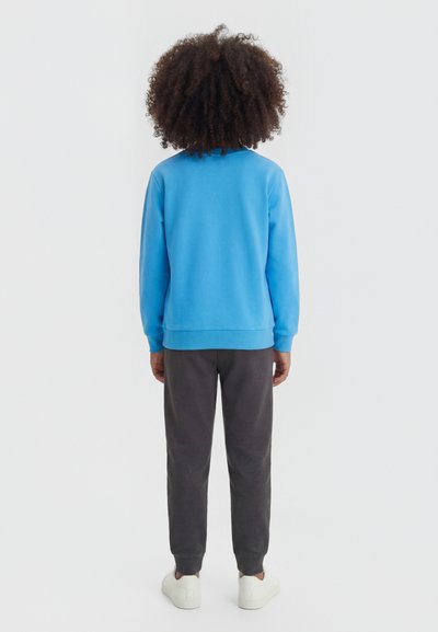 Enfant aux cheveux bouclés de dos, portant un sweat-shirt bleu, un pantalon foncé et des baskets blanches sur un fond uni.
