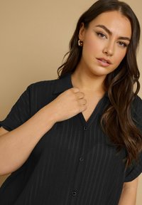Camicia nera con bottoni, caratterizzata da un motivo a righe testurizzato, con colletto a V e maniche corte. Il modello indossa orecchini a cerchio dorati.