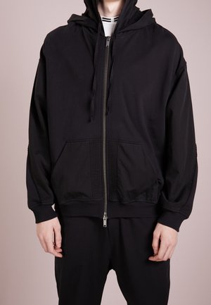 Black zip-up hoodie with a hood, featuring a textured panel on the front and two pockets. Made of soft fabric, relaxed fit.